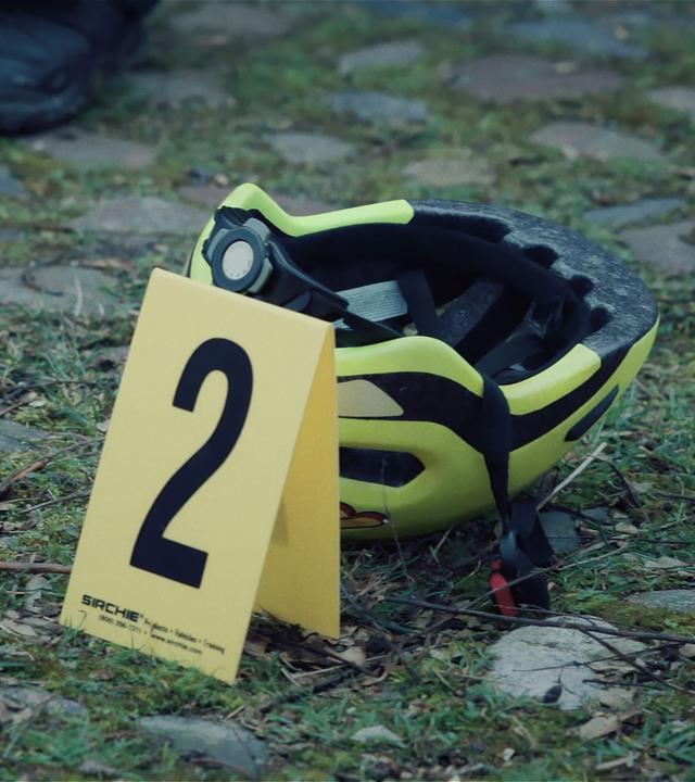 Auf einem mit grasbewachsenen Weg aus Kopfsteinplaster liegt ein neongelber Fahrradhelm. Daneben steht ein Aufsteller zum Kennzeichnen von Beweismitteln mit der Nummer 2 darauf.