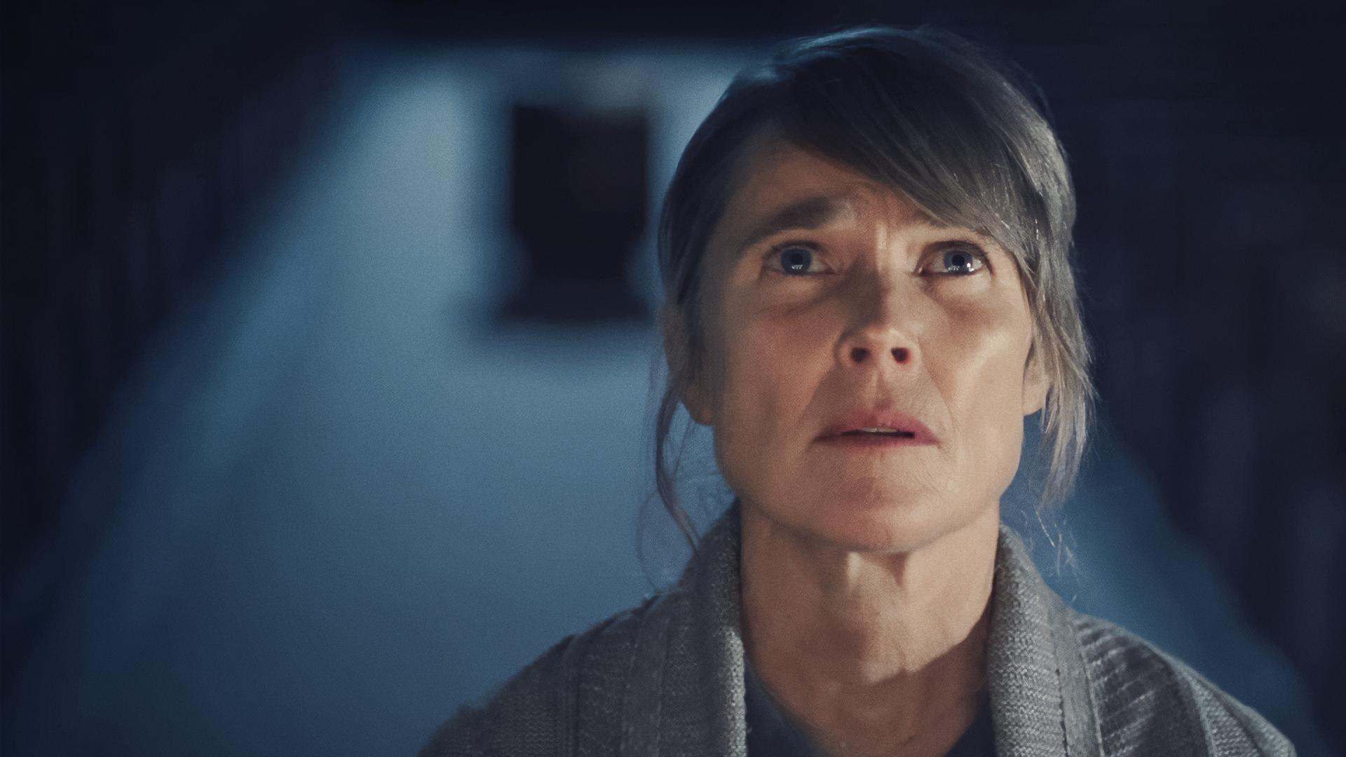 Maria Kerkel (Karoline Eichhhorn), eine blonde Frau mittleren Alters, die eine Kreuzkette um den Hals trägt, steht in einem dunklen Raum vor Kerzen und blickt mit bewegtem Gesichtsausdruck nach oben als würde sie beten.