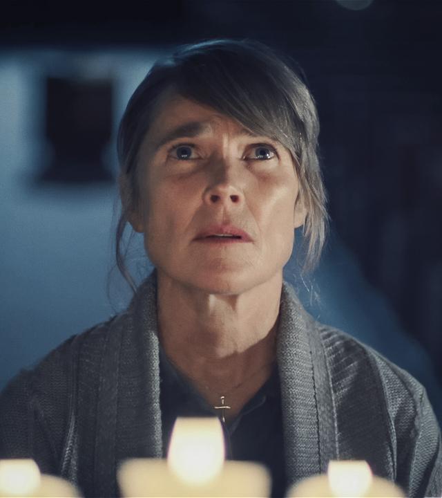 Maria Kerkel (Karoline Eichhhorn), eine blonde Frau mittleren Alters, die eine Kreuzkette um den Hals trägt, steht in einem dunklen Raum vor Kerzen und blickt mit bewegtem Gesichtsausdruck nach oben als würde sie beten.