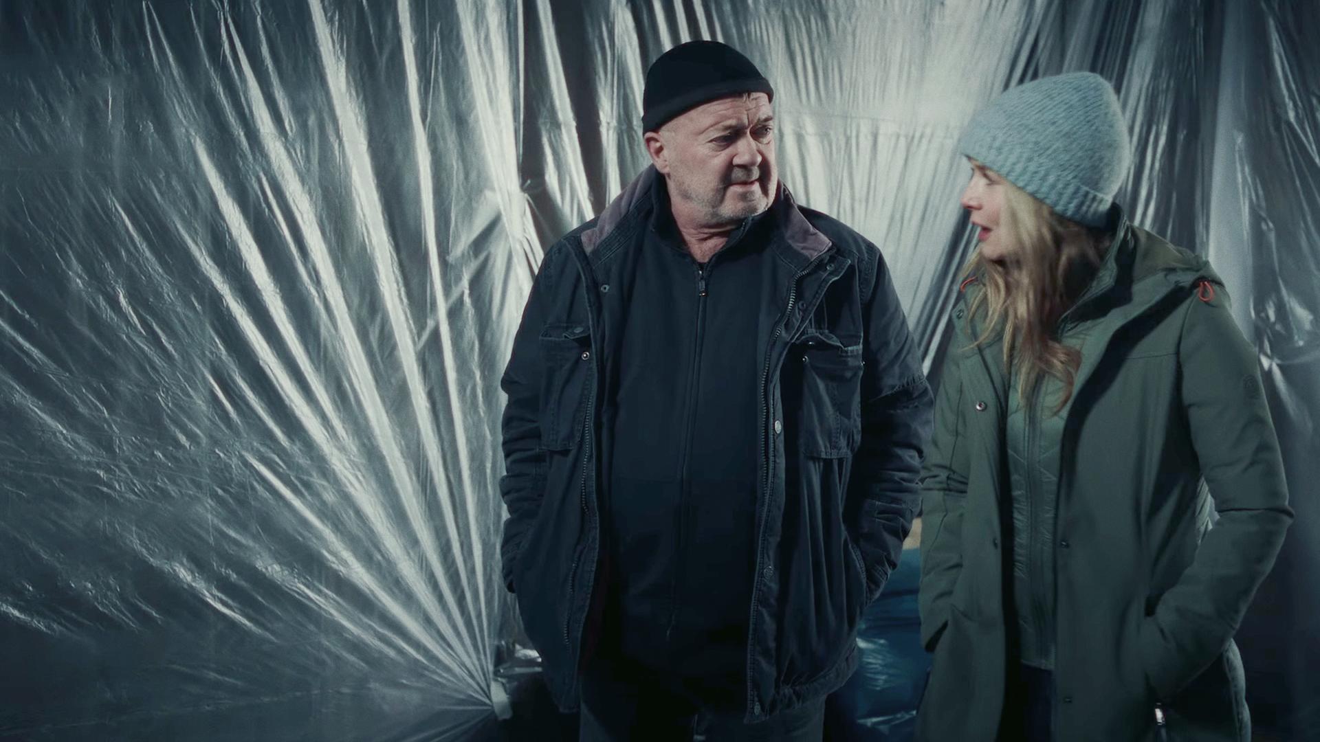 Die Kommissare Otto und Linett (Florian Martens, Stefanie Stappenbeck) stehen in Winterkleidung und Mützen vor einer Wand, die mit Plastikfolie verhängt ist.