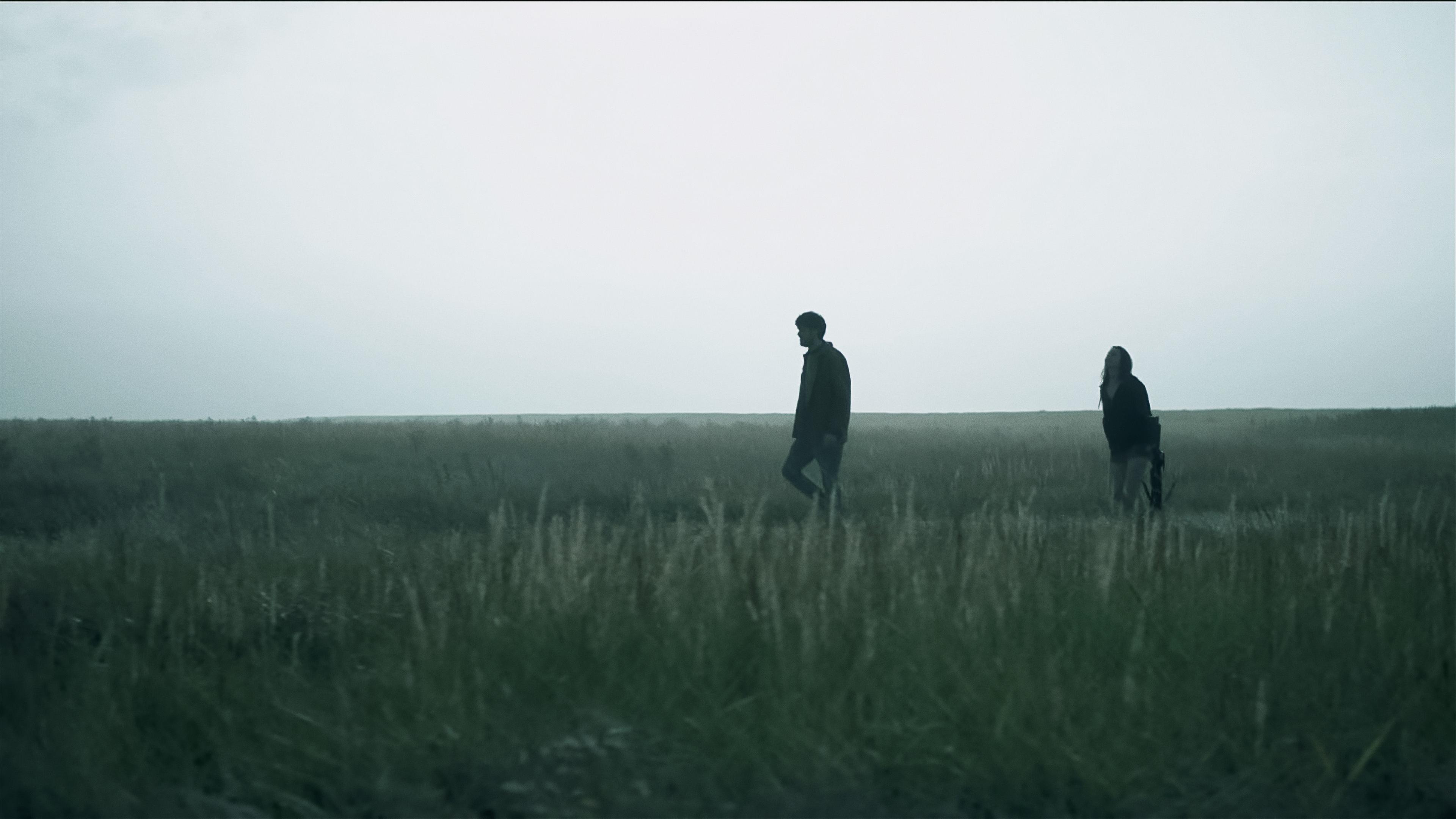 Ein Mann und eine Frau laufen bei düsterem Licht über eine Wiese. Die Frau trägt in der linken Hand eine Armbrust. 