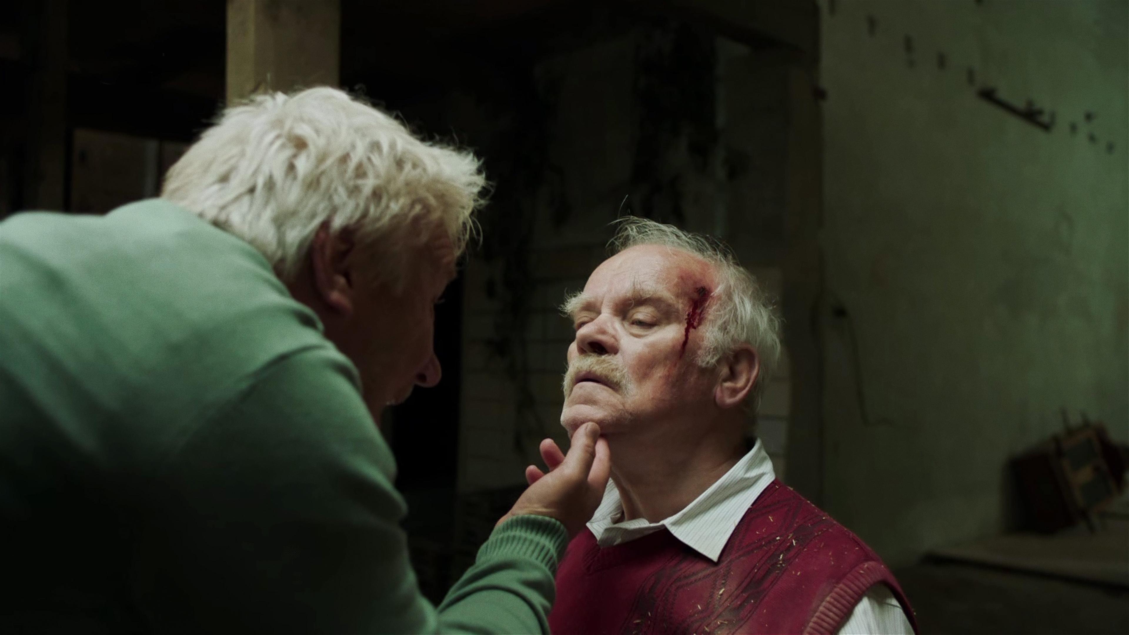 Heinrich Jansen (Peter Franke), ein alter Herr mit weißem Haar, Hemd und Pullunder sitzt einem anderen Mann gegenüber, dessen Gesicht man nicht sehen kann und der Jansens Kinn festhält. Jansens Schläe blutet.