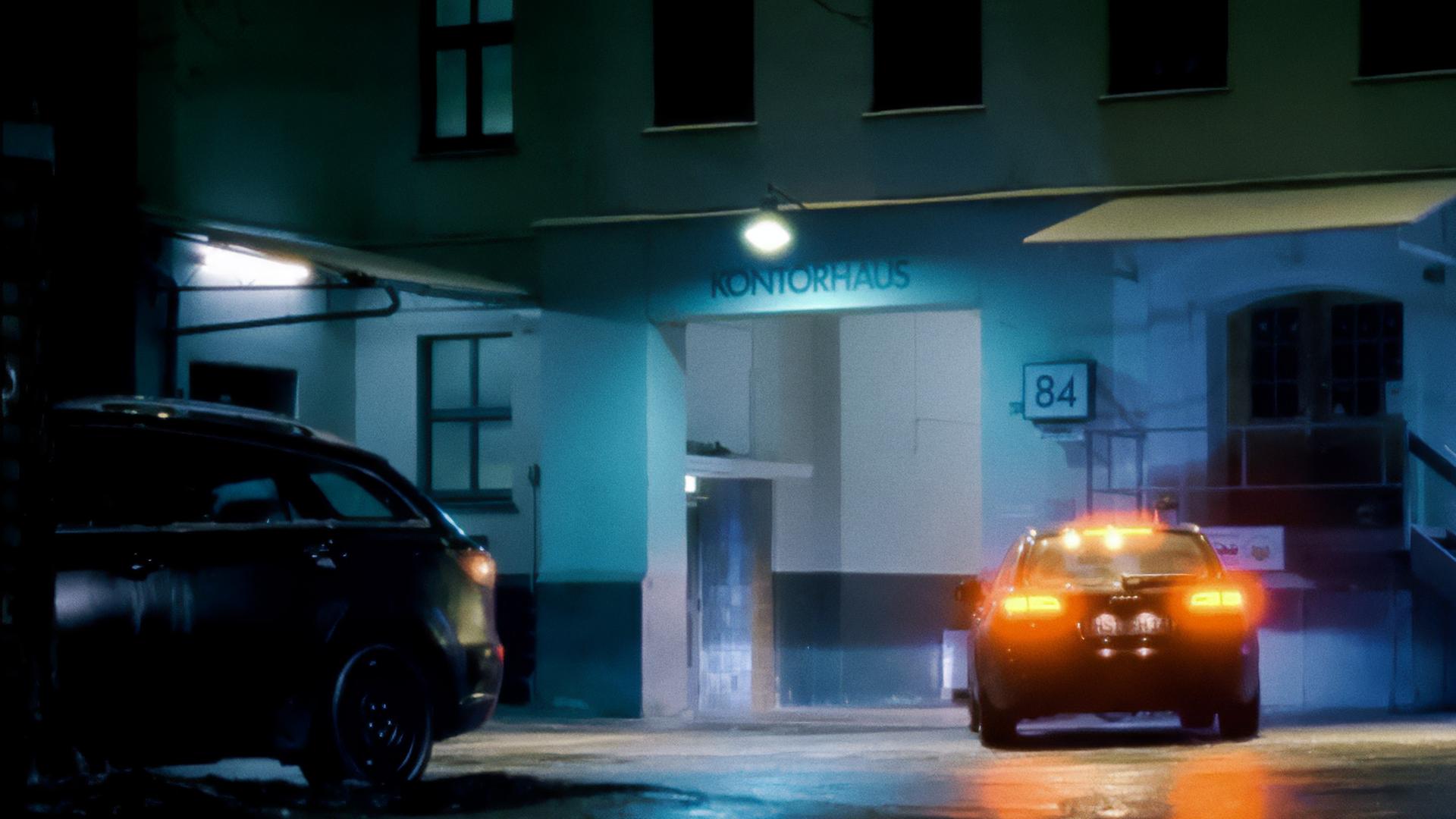 Ein Auto steht bei Nacht auf dem nassen Hof eines Gebäudekomplexes über dessen Eingang das Wort "Kontorhaus" steht. Die Rücklichter leuchten rot auf. 