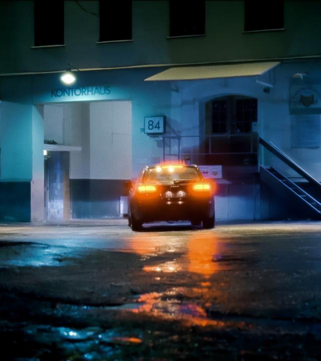Ein Auto steht bei Nacht auf dem nassen Hof eines Gebäudekomplexes über dessen Eingang das Wort "Kontorhaus" steht. Die Rücklichter leuchten rot auf. 