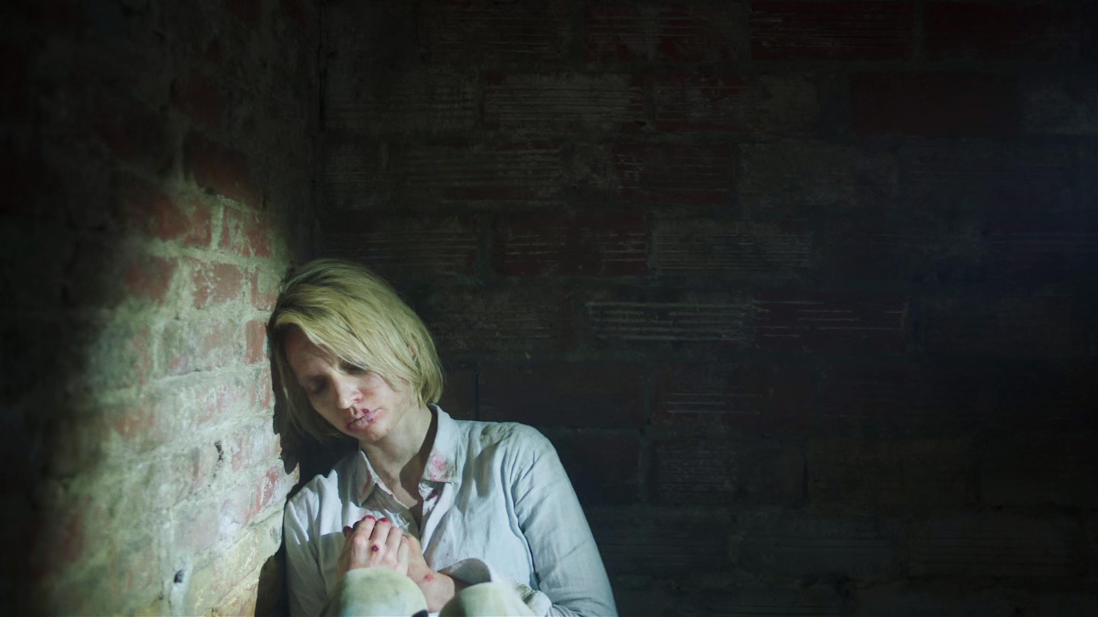 Judith (), eine blonde junge Frau, sitzt in sich zusammengesackt in der Ecke eines dunklen Raums mit Backsteinwänden. Im Gesicht, an den Händen und an ihrer Bluse ist Blut.