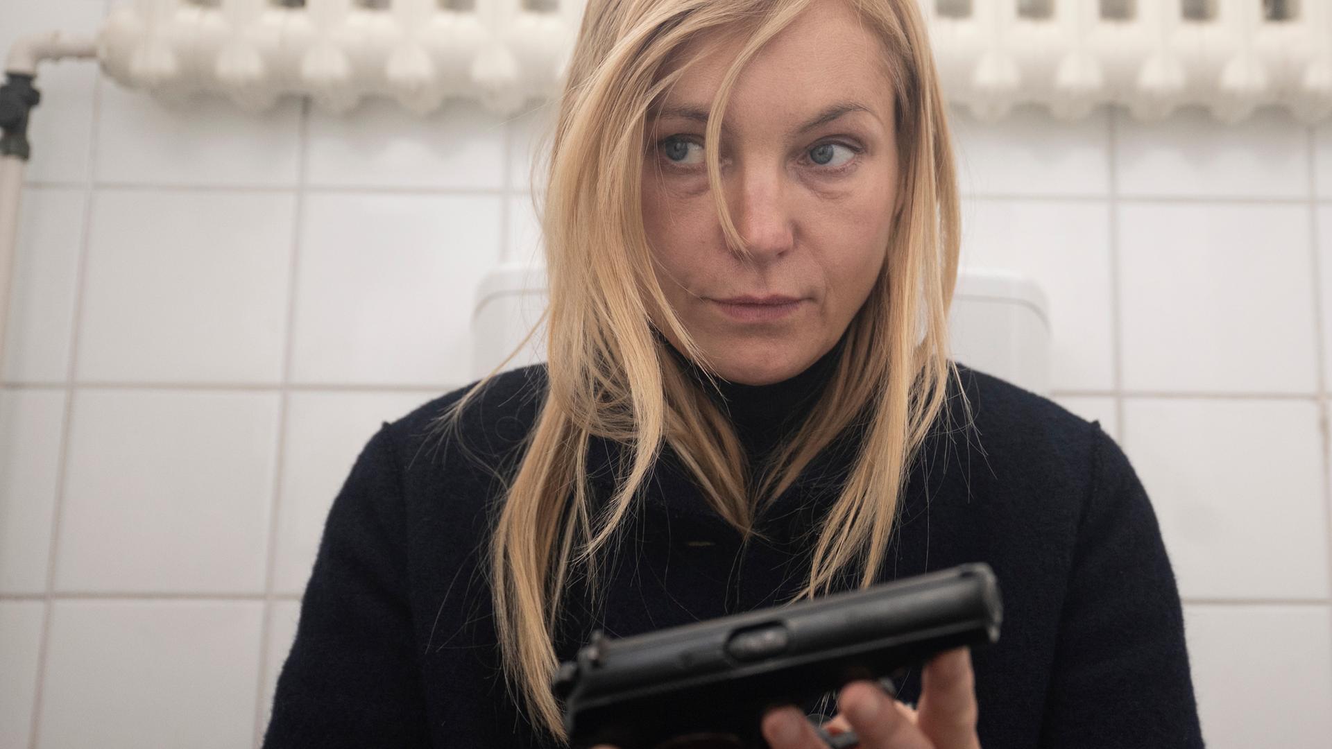 Judith Schrader (Nadja Uhl) sitzt allein in der WC-Kabine mit einer Waffe in der Hand, ihr Blick wirkt nachdenklich und angespannt.