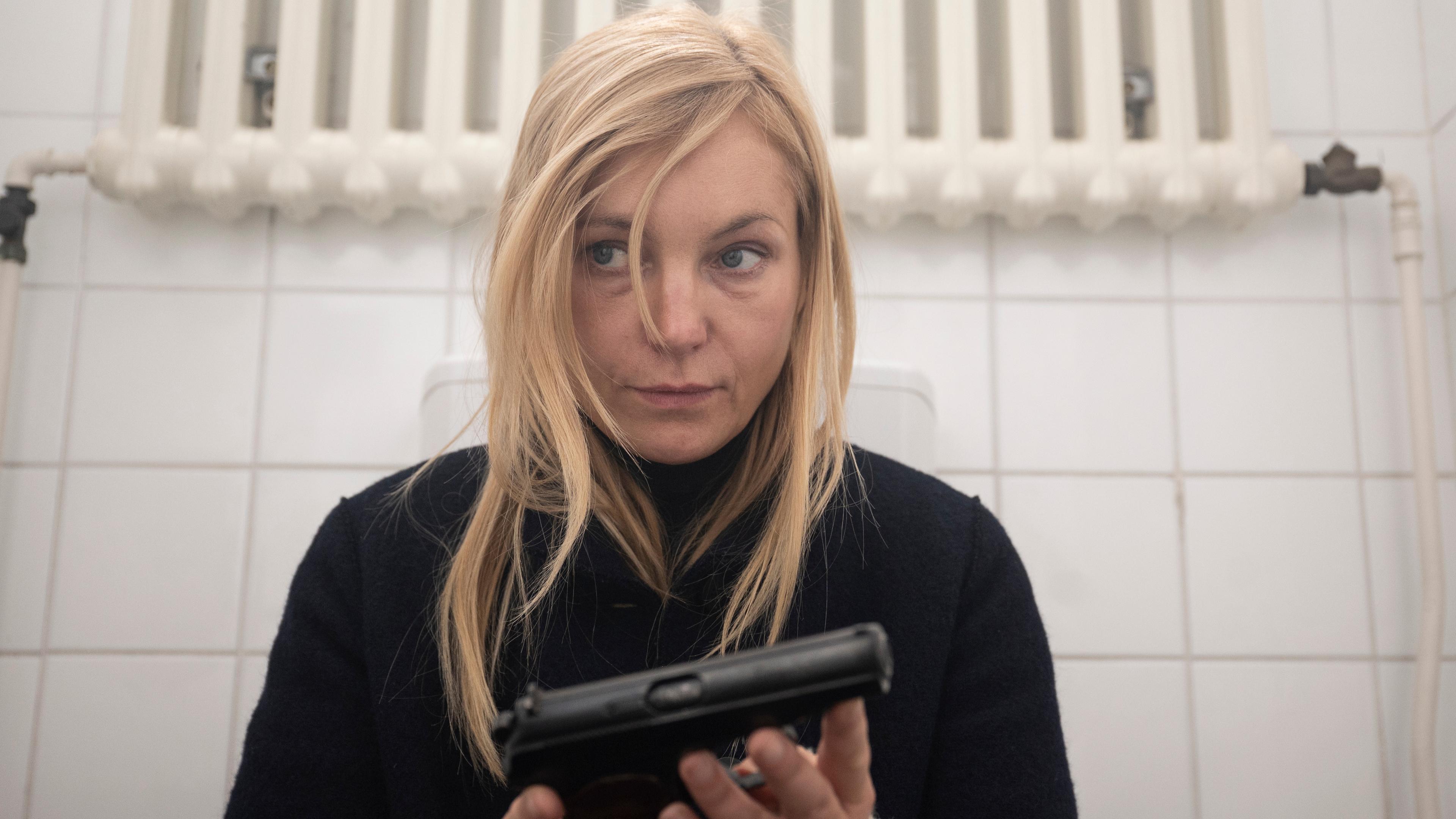 Judith Schrader (Nadja Uhl) sitzt allein in der WC-Kabine mit einer Waffe in der Hand, ihr Blick wirkt nachdenklich und angespannt.
