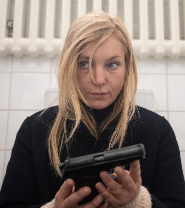 Judith Schrader (Nadja Uhl) sitzt allein in der WC-Kabine mit einer Waffe in der Hand, ihr Blick wirkt nachdenklich und angespannt.