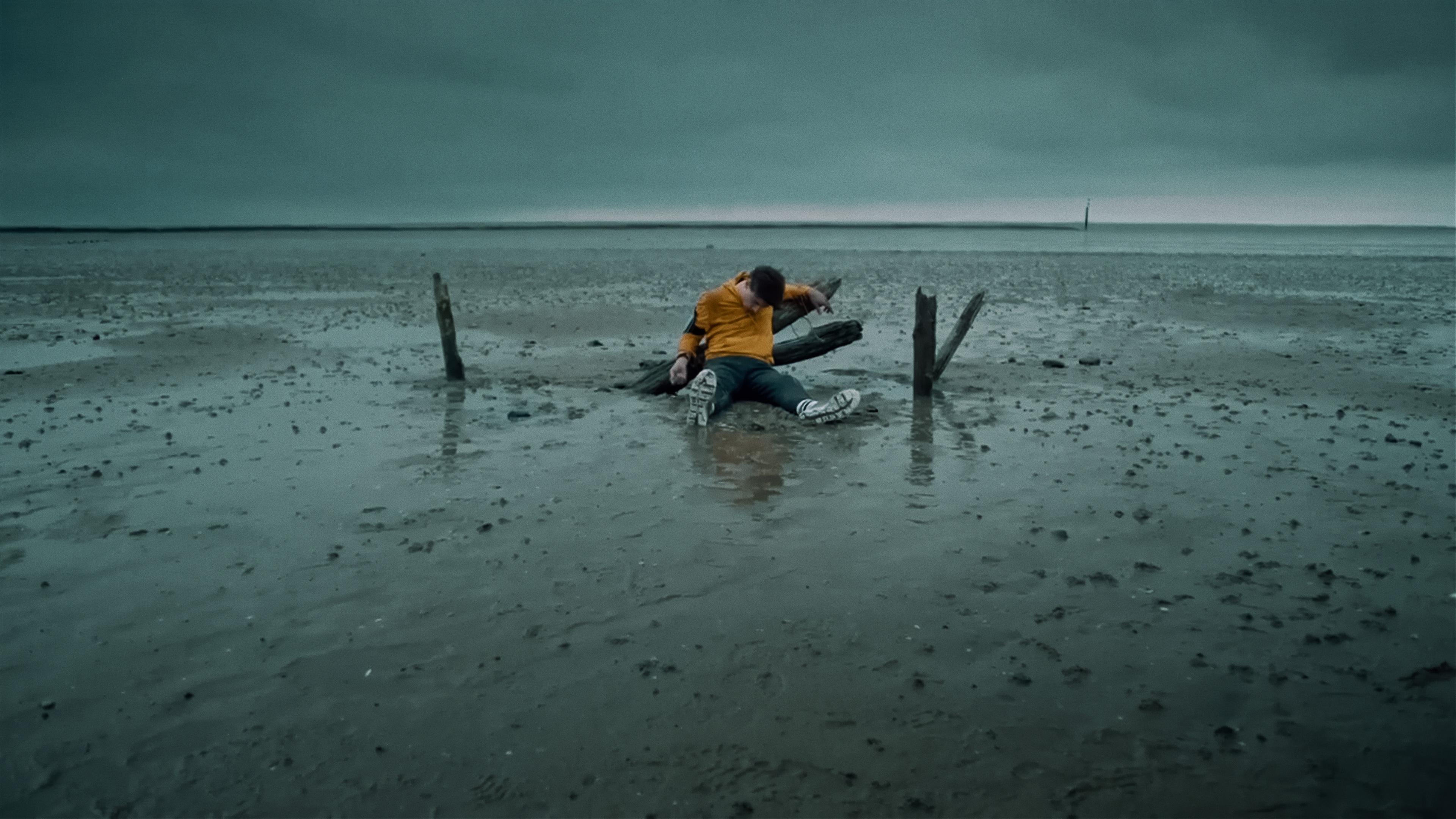 Im Watt liegt eine Leiche; die Hände sind abgebrochene Pfähle gebunden, die aus dem Sand ragen. 