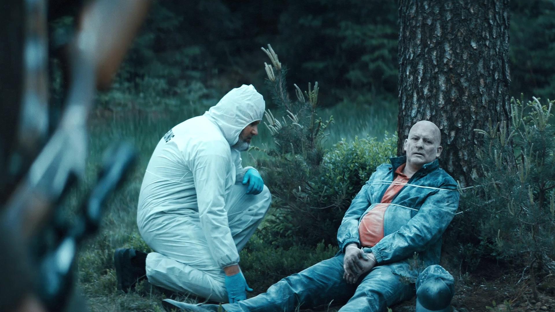 Die tiefgerorene und mit Frost besetzte Leiche eines Mannes sitzt an einen Baum gefesselt auf einer Wiese, während sich ein Mann von der Spurensicherung in einem weißen Schutzanzug neben ihm kniet. 