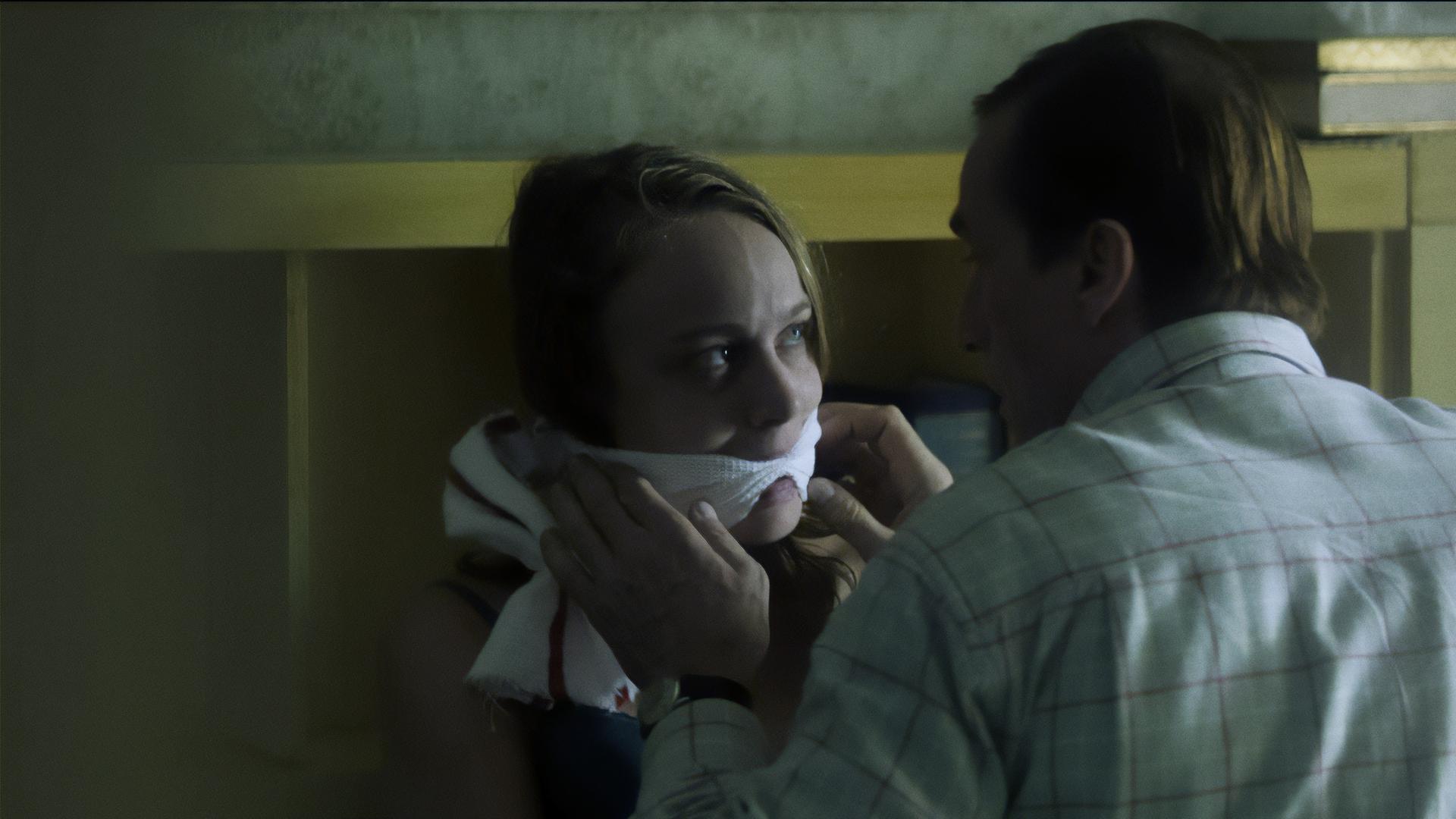 Ein Mann kniet in einem Zimmer vor einer geknebelten Frau und richtet das Handtuch, das um ihren Mund gewickelt ist. 