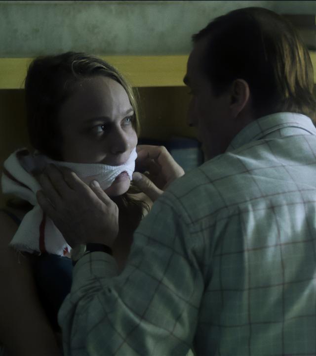 Ein Mann kniet in einem Zimmer vor einer geknebelten Frau und richtet das Handtuch, das um ihren Mund gewickelt ist. 