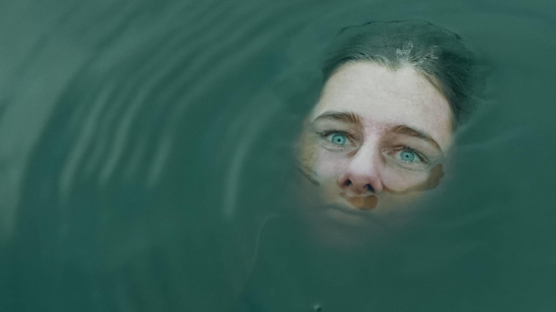 Das Gesicht einer Frau ragt teilweise aus trübem Wasser hervor. Ihre Augen sehen starr in die Ferne. 