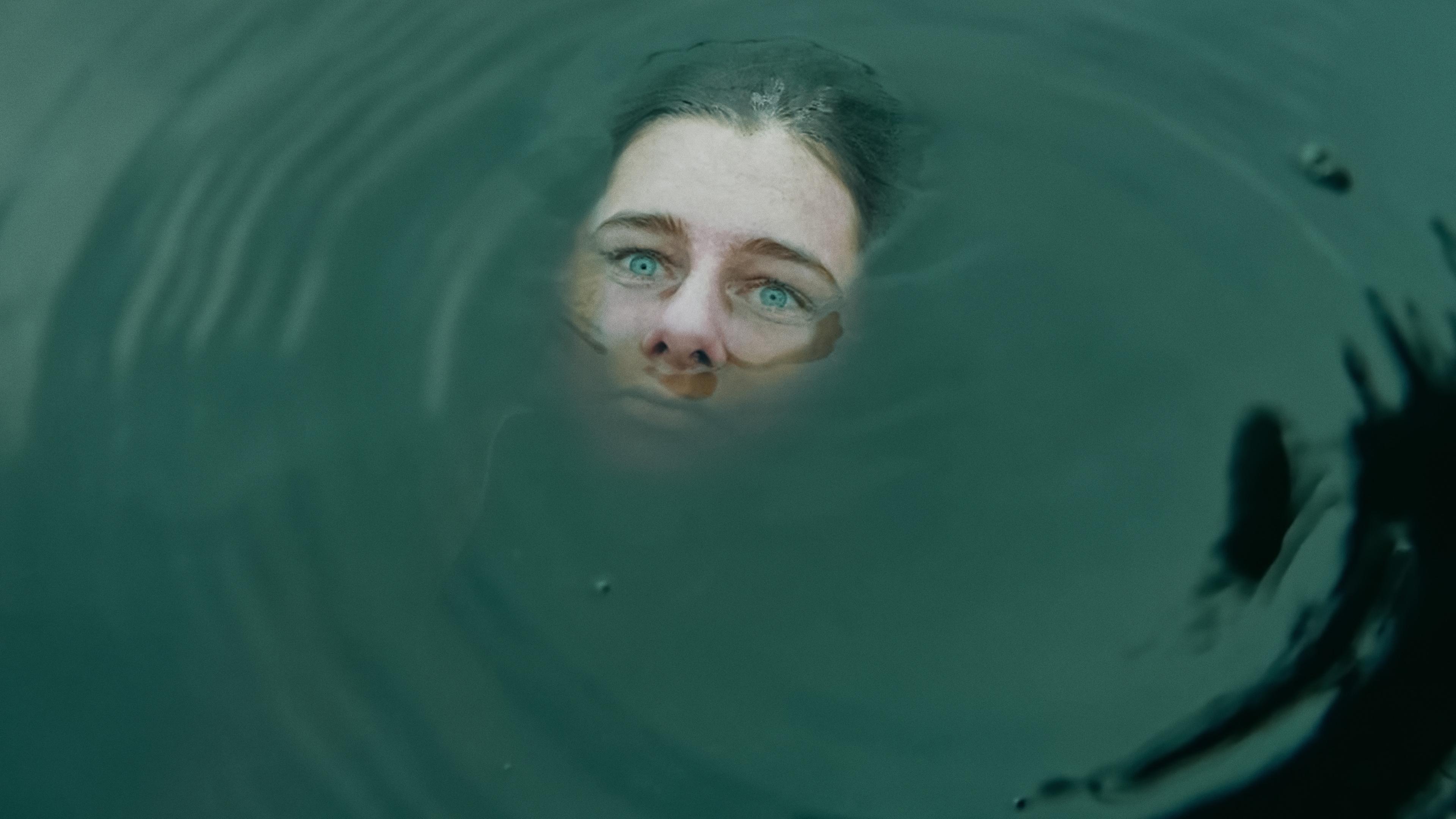 Das Gesicht einer Frau ragt teilweise aus trübem Wasser hervor. Ihre Augen sehen starr in die Ferne. 