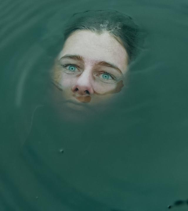 Das Gesicht einer Frau ragt teilweise aus trübem Wasser hervor. Ihre Augen sehen starr in die Ferne. 
