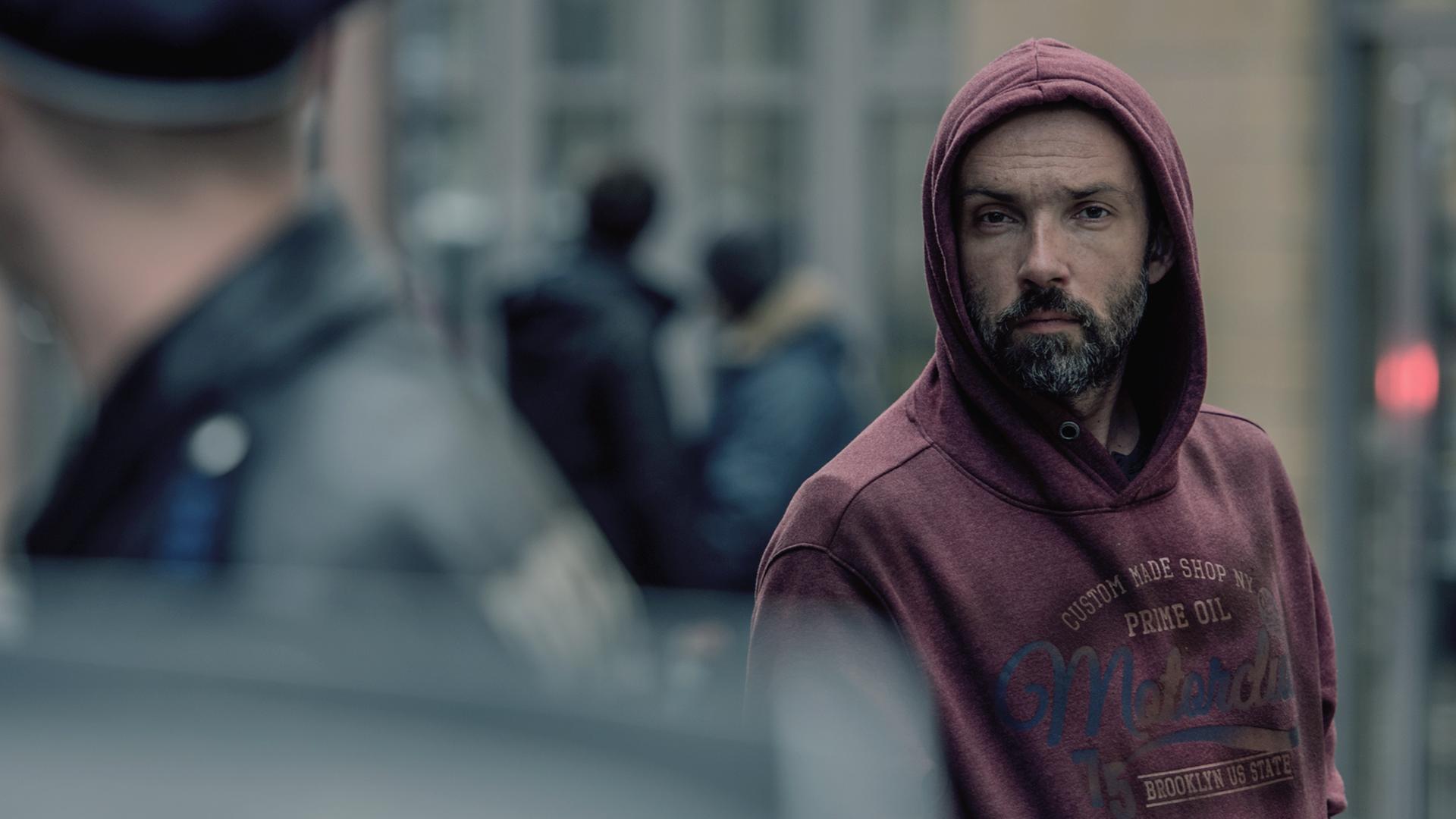 Anton Robak (Lucas Gregorowicz), ein Mann mit Bart und einem roten Kaputzenpulli, beobachtet einen Polizisten, der im Vordergrund des Bildes nur verschwommen zu erkennen ist.