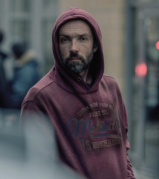 Anton Robak (Lucas Gregorowicz), ein Mann mit Bart und einem roten Kaputzenpulli, beobachtet einen Polizisten, der im Vordergrund des Bildes nur verschwommen zu erkennen ist.