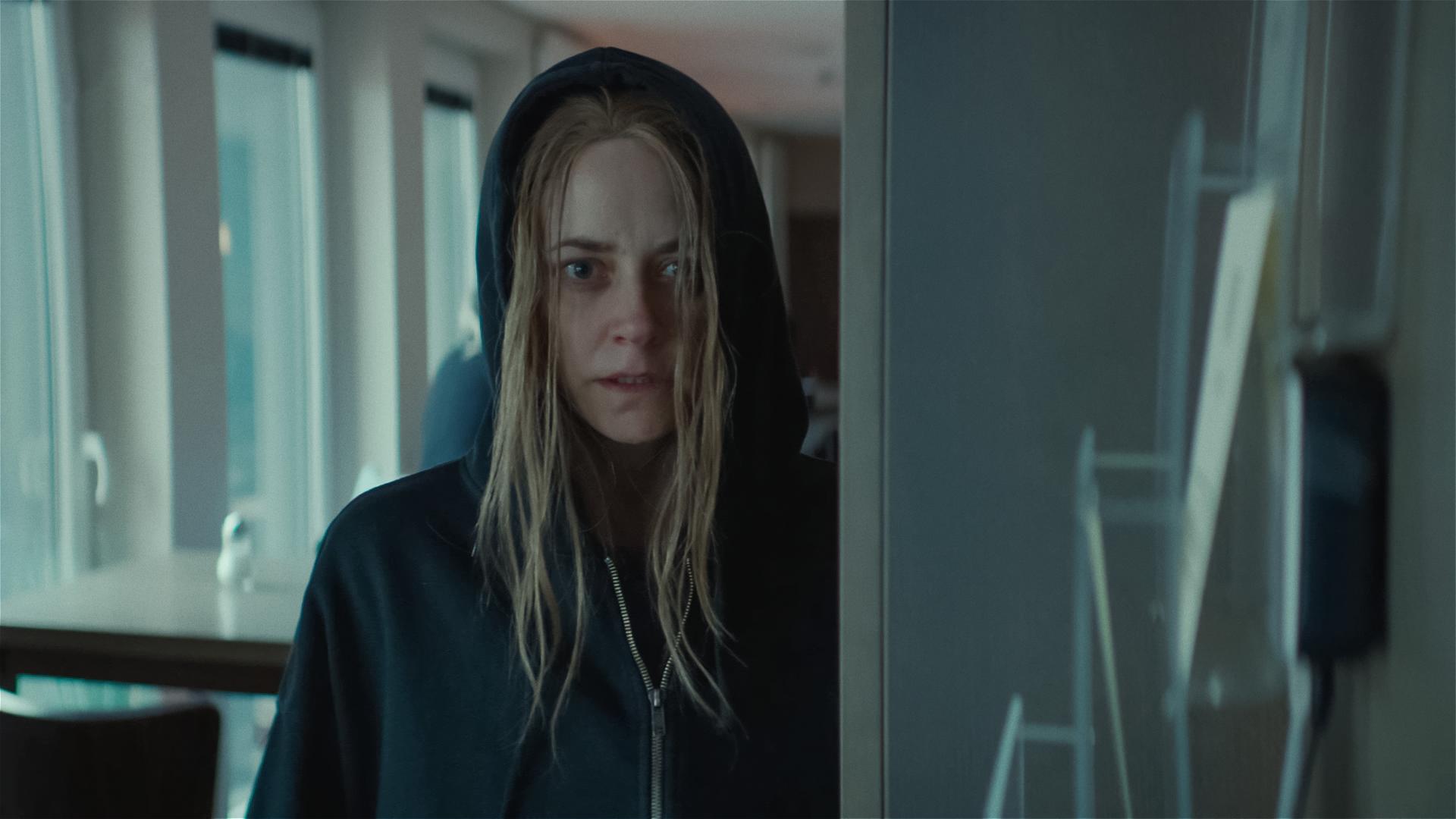 Svenja Moers (Anja Knauer), eine junge Frau mit strähnigen blonden Haaren, steht in einem Raum und scheint etwas zu beobachten. Sie trägt ein schwarzes Sweatshirt und hat sie die Kapuze übergezogen.