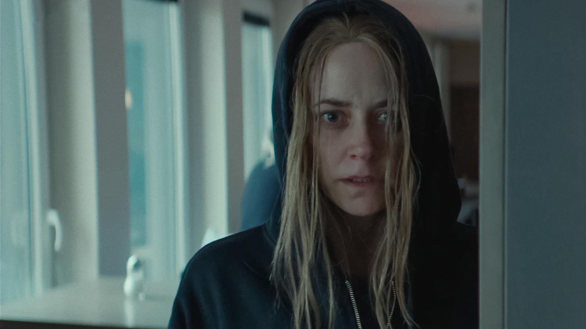 Svenja Moers (Anja Knauer), eine junge Frau mit strähnigen blonden Haaren, steht in einem Raum und scheint etwas zu beobachten. Sie trägt ein schwarzes Sweatshirt und hat sie die Kapuze übergezogen.