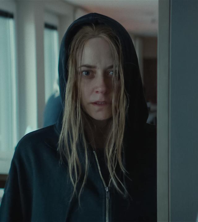 Svenja Moers (Anja Knauer), eine junge Frau mit strähnigen blonden Haaren, steht in einem Raum und scheint etwas zu beobachten. Sie trägt ein schwarzes Sweatshirt und hat sie die Kapuze übergezogen.