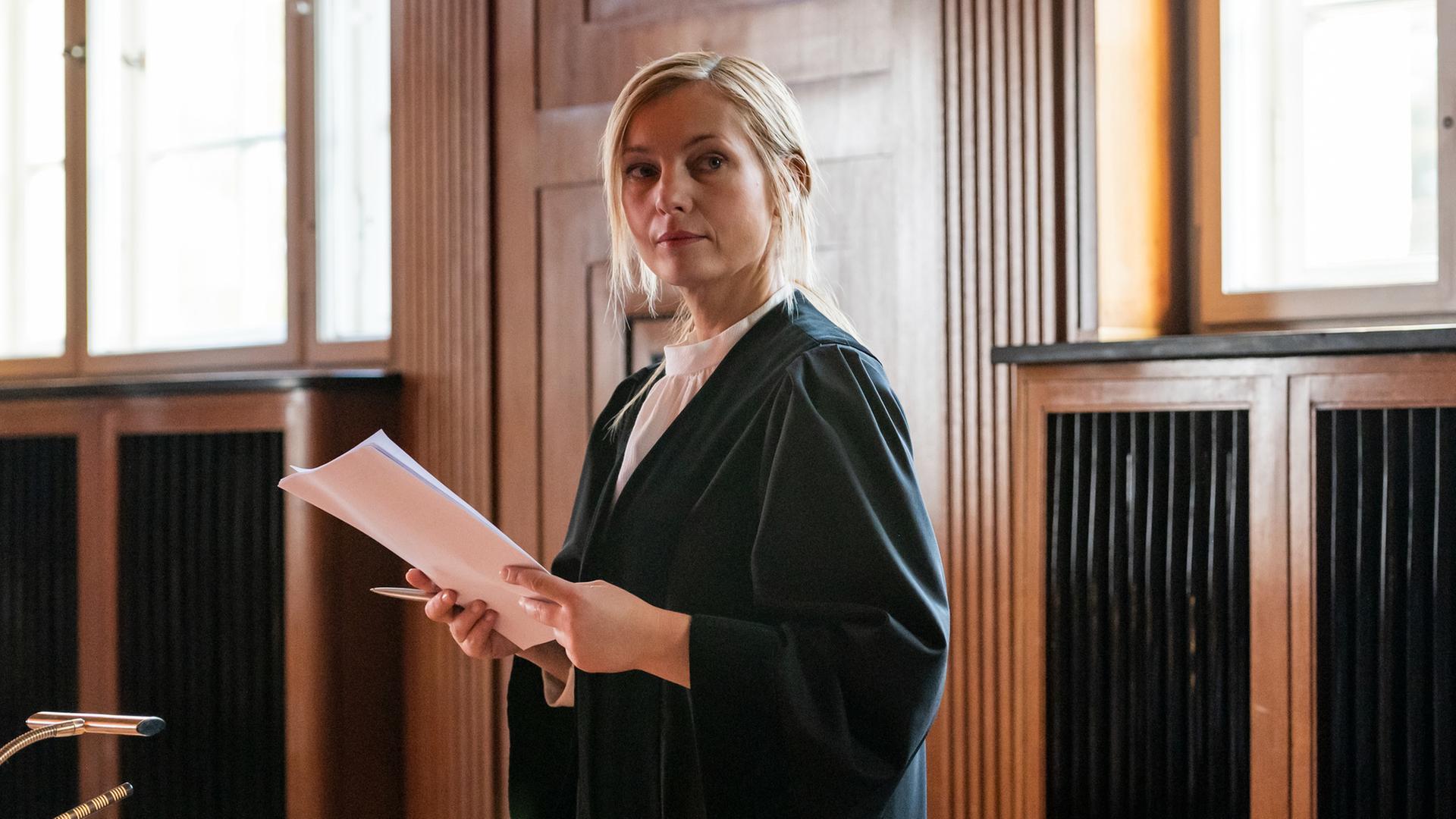 Staatsanwältin Judith Schrader (Nadja Uhl) in ihrer Anwaltsrobe im Verhandungssaal des Gerichtsgebäudes. In der Hand hält sie einen Stoß Dokumente.