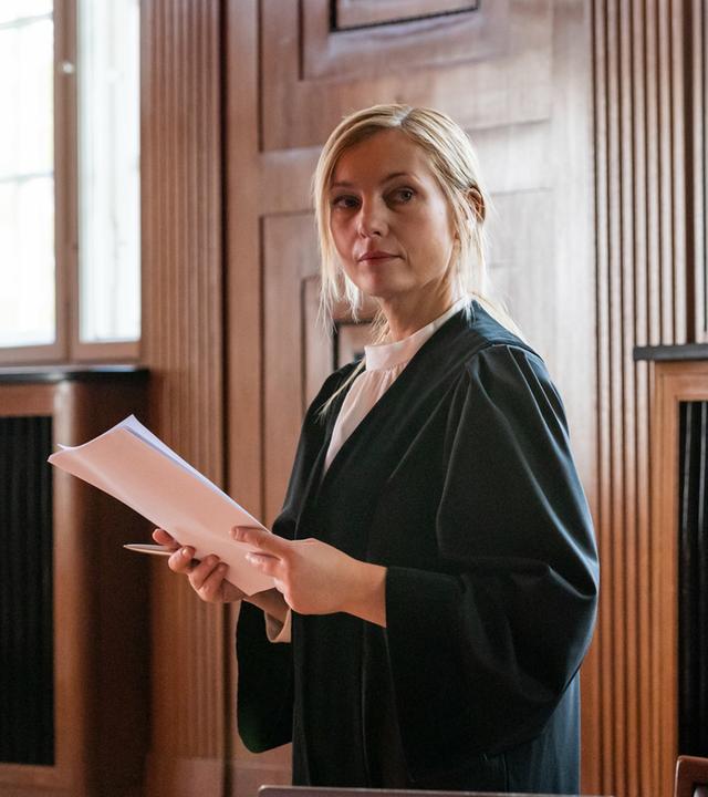 Staatsanwältin Judith Schrader (Nadja Uhl) in ihrer Anwaltsrobe im Verhandungssaal des Gerichtsgebäudes. In der Hand hält sie einen Stoß Dokumente.