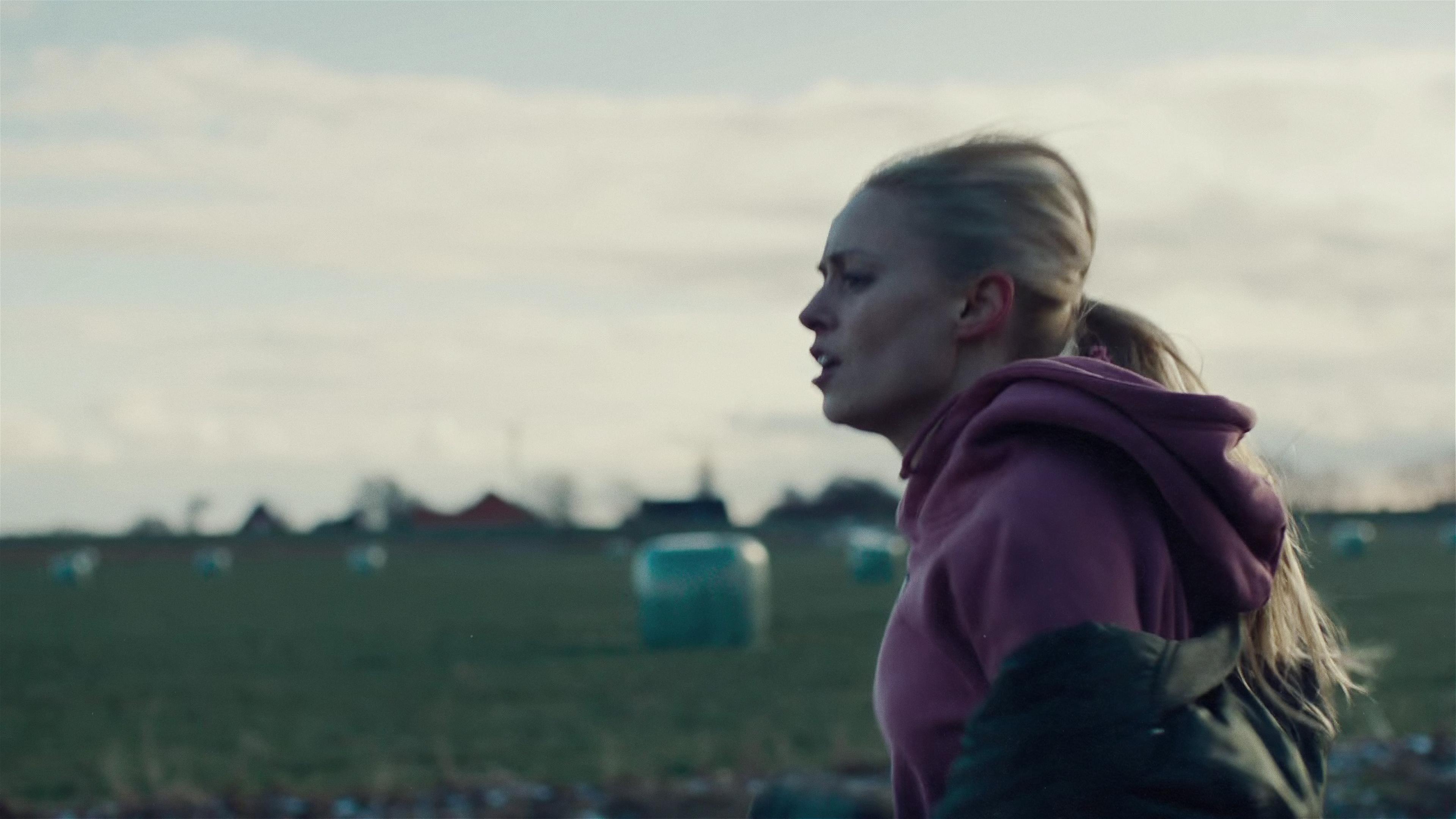 Eine blonde, junge Frau mit Pferdeschwanz, rosa Sweatshirt und dunklem Anorack läuft auf einem Feld vor etwas oder jemandem davon.