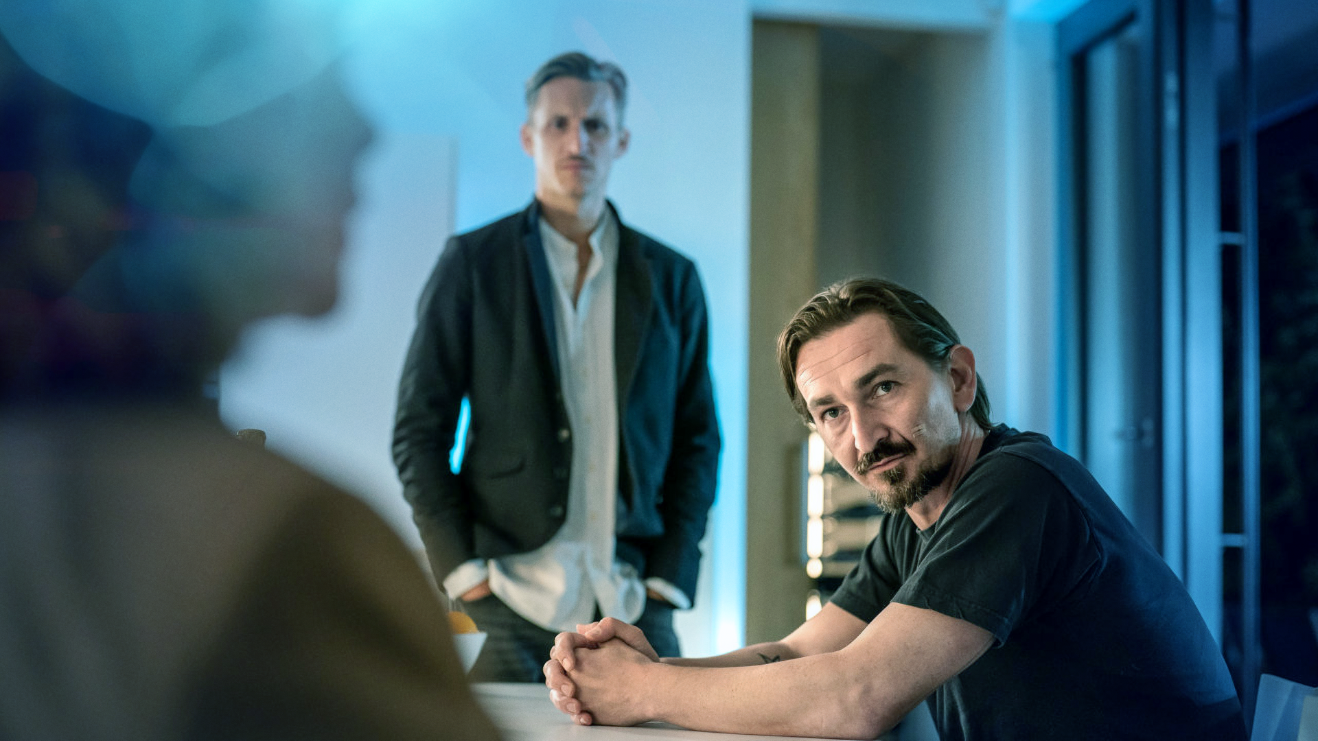 An einem Tisch sitzt ein Mann. Im Hintergrund steht Moritz Brenner (Johannes Hendrik Langer). Sie unterhalten sich mit einer Frau, die nur von hinten zu sehen ist. 