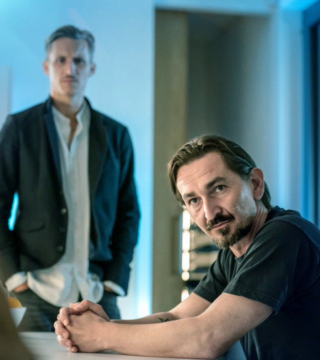 An einem Tisch sitzt ein Mann. Im Hintergrund steht Moritz Brenner (Johannes Hendrik Langer). Sie unterhalten sich mit einer Frau, die nur von hinten zu sehen ist. 