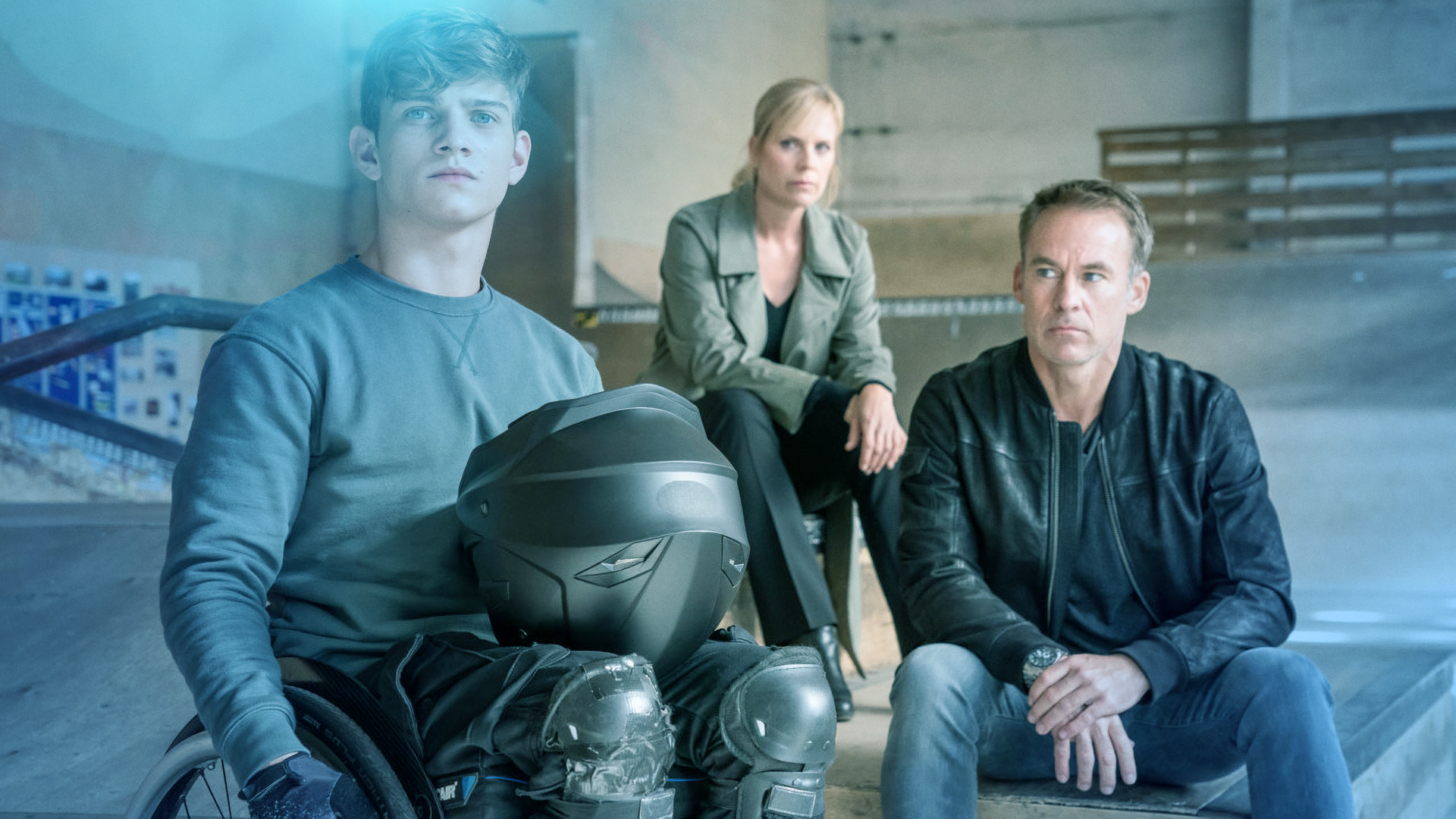 Ein junger Mann im Rollstuhl mit Knieschonern und Motorradhelm auf dem Schoß. Im Hintergrund Ina Zimmermann (Melanie Marschke) und Jan Maybach (Marco Girnth)