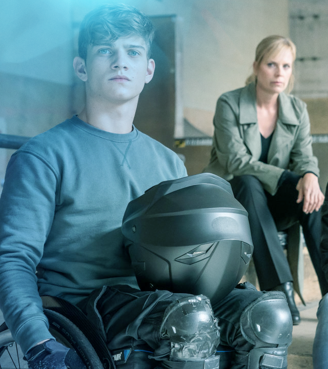 Ein junger Mann im Rollstuhl mit Knieschonern und Motorradhelm auf dem Schoß. Im Hintergrund Ina Zimmermann (Melanie Marschke) und Jan Maybach (Marco Girnth)