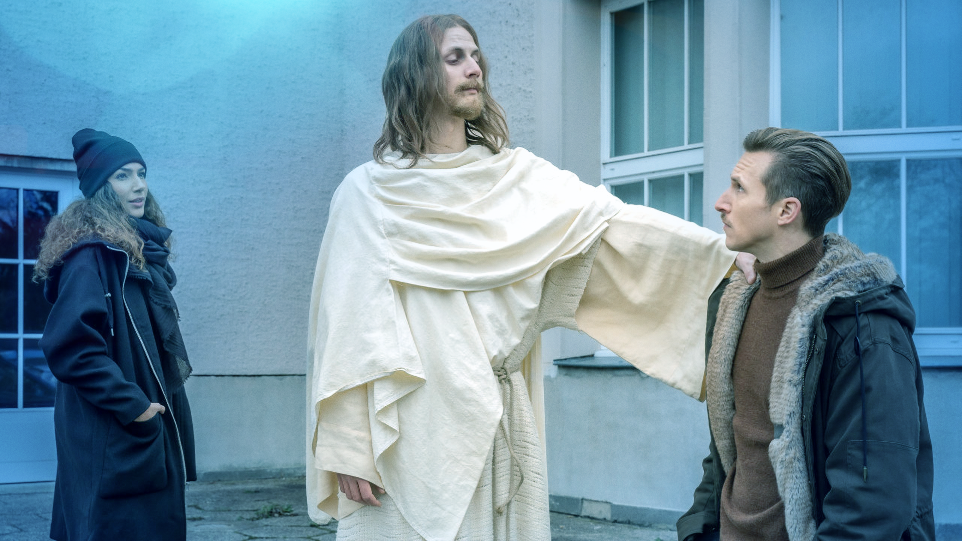 Ein Mann in einem Jesus-Kostüm legt Moritz Brenner (Johannes Hendrik Langer) die Hand auf die Schulter während Kim Nowak (Amy Mußul) zusieht. 