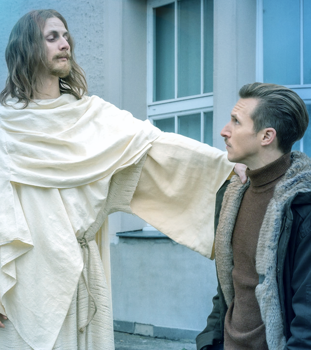 Ein Mann in einem Jesus-Kostüm legt Moritz Brenner (Johannes Hendrik Langer) die Hand auf die Schulter während Kim Nowak (Amy Mußul) zusieht. 