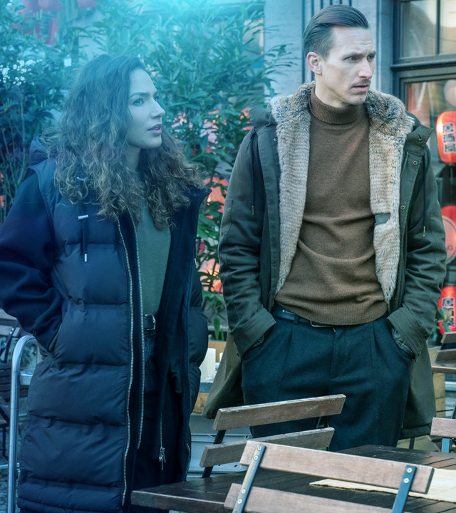 Auf der Terrasse eines Asia-Restaurants stehen Kim Novak (Amy Mußul) und Moritz Brenner (Johannes Hendrik Langer) neben einem Koch, der Babus-Tischsets in der Hand hält. 