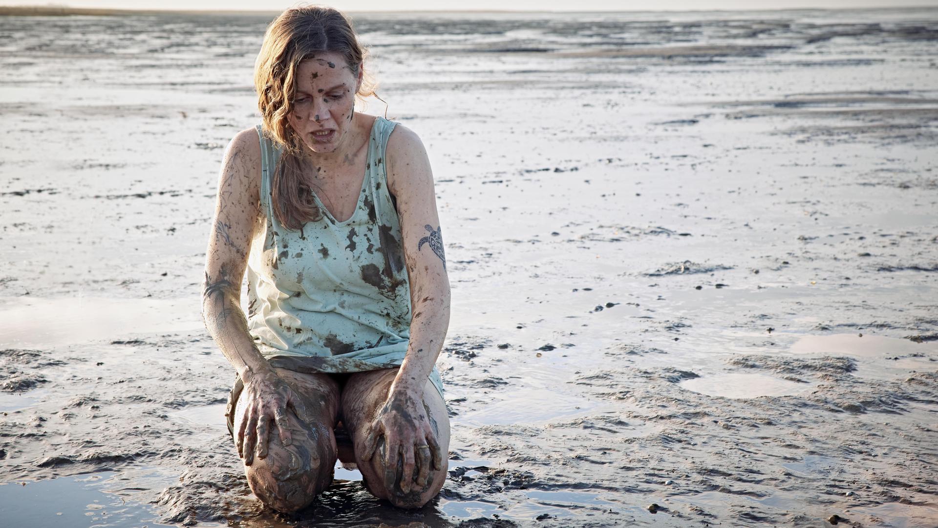 Ann Kathrin Klaasen (Picco von Groote) kniet im Watt nur mit einem Hemd bekleidet. Sie ist über und über mit Schlamm verschmiert. Sie schaut entsetzt auf ihre Knie: Überall auf ihrem Körper sind Tattoos.