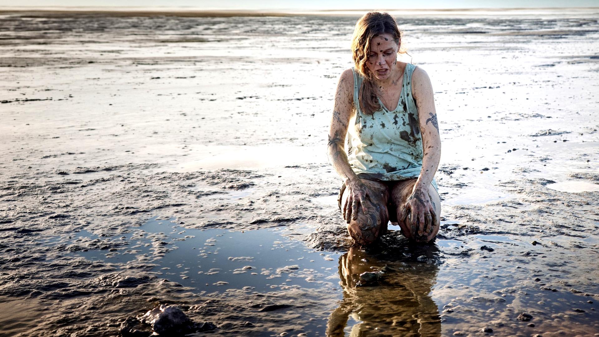 Ann Kathrin Klaasen (Picco von Groote) kniet im Watt nur mit einem Hemd bekleidet. Sie ist über und über mit Schlamm verschmiert. Sie schaut entsetzt auf ihre Knie: Überall auf ihrem Körper sind Tattoos.