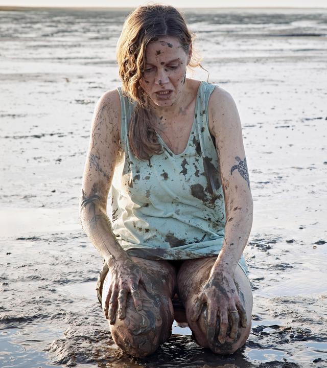 Ann Kathrin Klaasen (Picco von Groote) kniet im Watt nur mit einem Hemd bekleidet. Sie ist über und über mit Schlamm verschmiert. Sie schaut entsetzt auf ihre Knie: Überall auf ihrem Körper sind Tattoos.