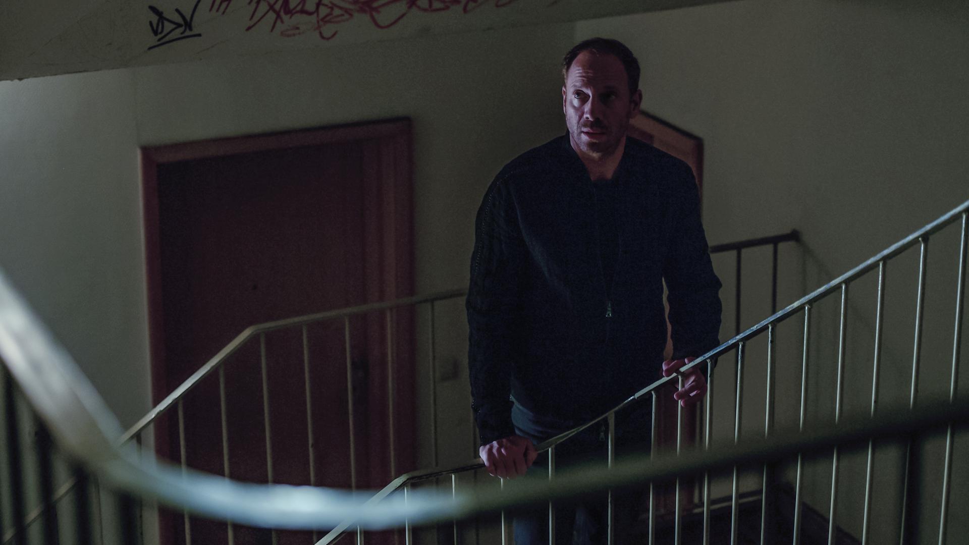 Professor Oliver Lauder (Johann von Bülow), ein Mann in einer schwarzen Jacke, steht in einem spärlich beleuchteten Treppenhaus.