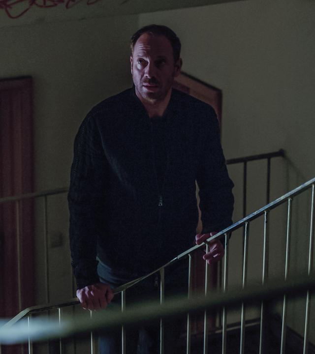 Professor Oliver Lauder (Johann von Bülow), ein Mann in einer schwarzen Jacke, steht in einem spärlich beleuchteten Treppenhaus.