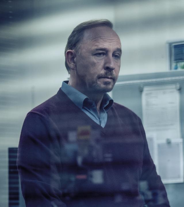 Karl Hidde (Alexander Held) steht nachdenklich im Büro. 