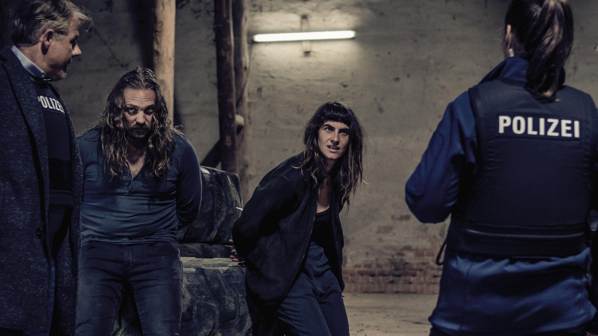 Hanne Beckmann (Samia Chancrin) und Peter Gruber (Denis Schmidt) stehen in einer Scheune. Vor ihnen zwei Polizisten, die nur von hinten zu sehen sind. 