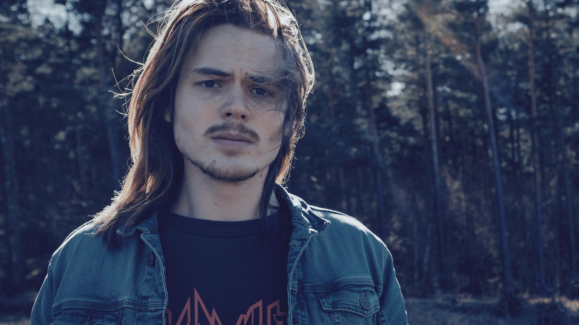 Jo Brand (Jack Owen Berglund), ein junger Mann mit langen Haaren und in einer Jeansjacke, steht vor einem Wald und schuat besorgt.