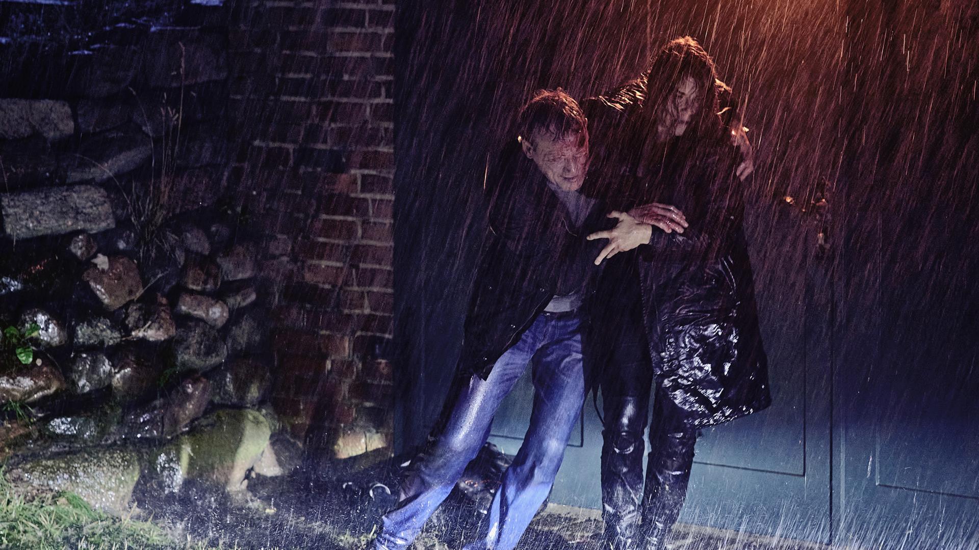 Nina Petersen (Katharina Wackernagel) stützt den am Kopf blutenden Benjamin Lietz (Wotan Wilke Möhring) auf dem Weg zum Auto. Es ist nacht und regnet stark.