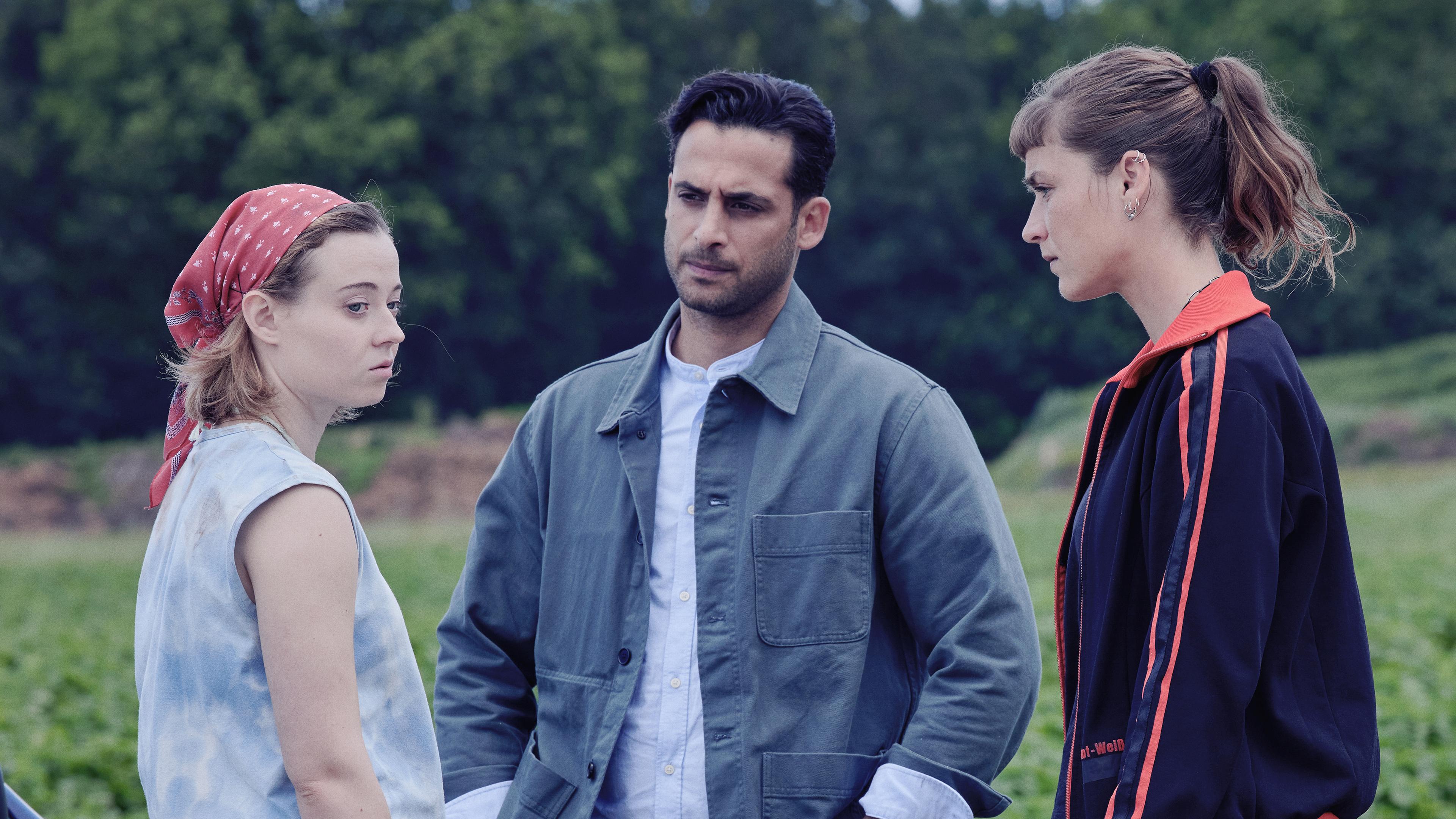 Jule Zabek (Sophie Pfennigstorf) und Karim Uthman (Karim Günes) befragen Blanka Wozniak (Linda Belinda Podszus) zu dem Toten auf der Landstraße. Sie stehen neben einem blauen Auto im Freien.