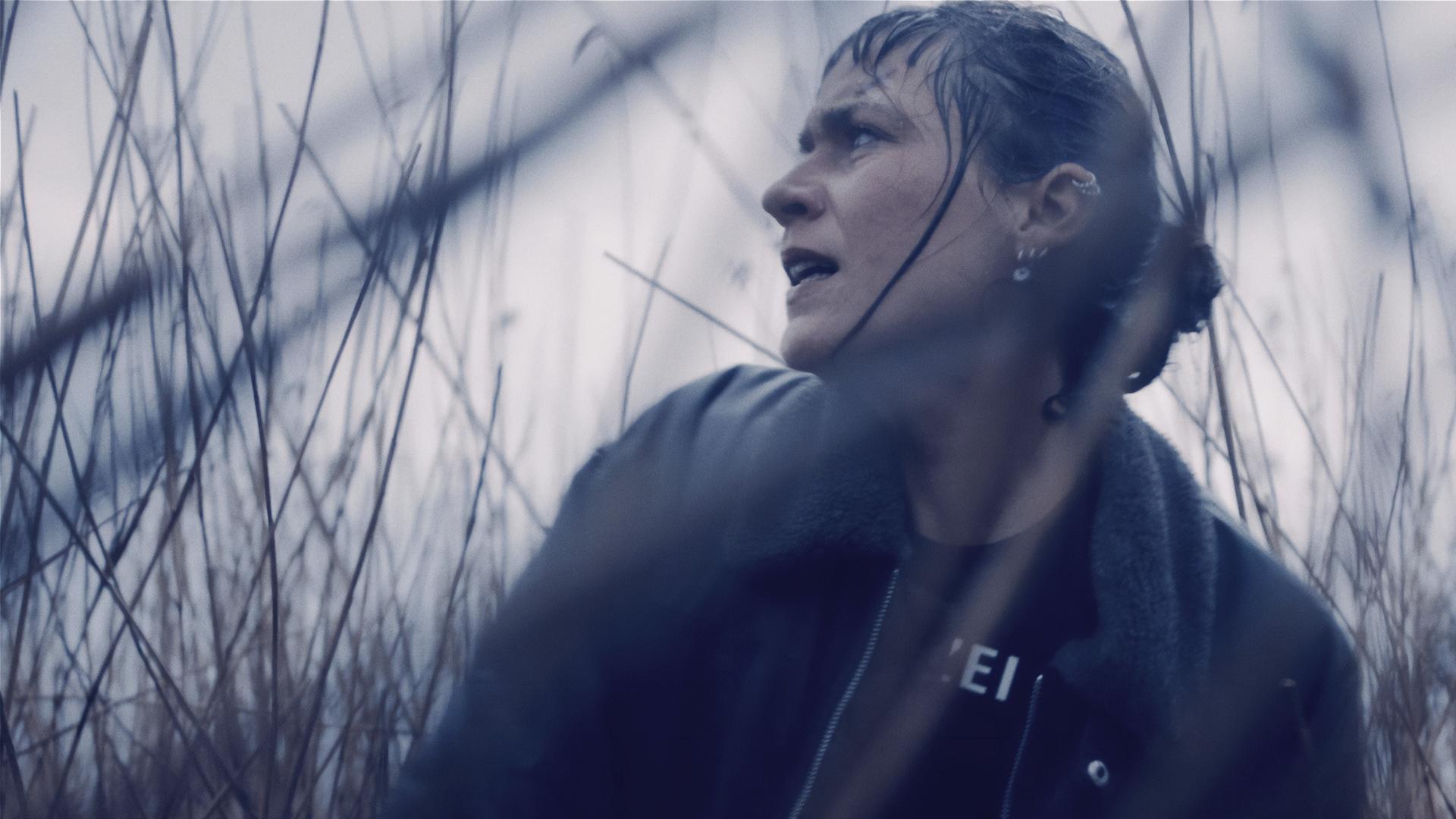 Jule Zabek (Sophie Pfennigstorf), eine junge Frau mit Pferdeschwanz und vielen Piercings im Ohr, kauert im hohen Gras und sieht sich über ihre Schulter nach etwas oder jemandem um. Sie ist sehr nassgeregnet.