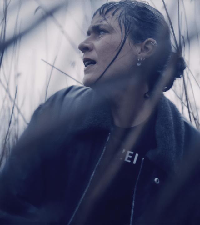 Jule Zabek (Sophie Pfennigstorf), eine junge Frau mit Pferdeschwanz und vielen Piercings im Ohr, kauert im hohen Gras und sieht sich über ihre Schulter nach etwas oder jemandem um. Sie ist sehr nassgeregnet.