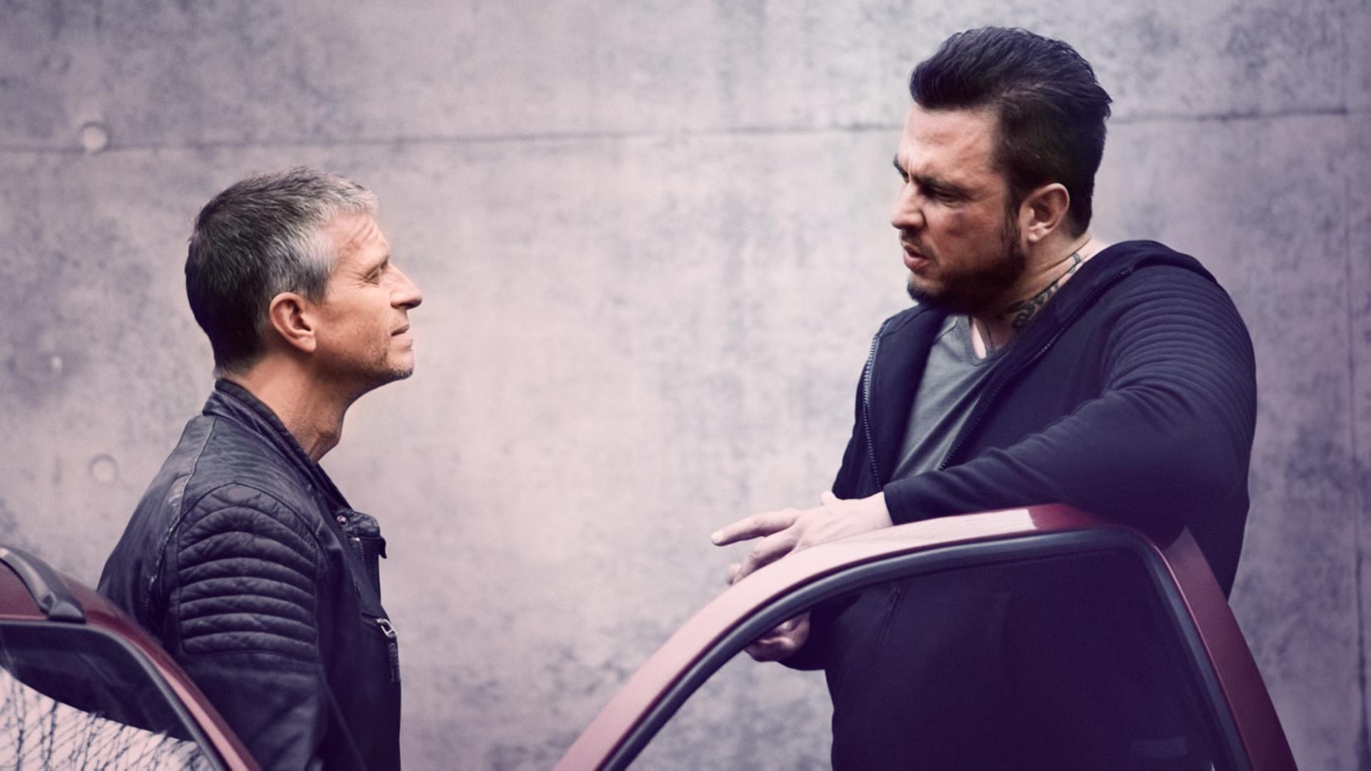 Peter (Dirk Heinrichs), ein Man mit einer schwarzen Lederjacke, blickt zu Dennis (Adrian Lynn) hoch, ein sehr großer und breiter Mann mit einem Tattoo am Hals. Die beiden stehen an einem Auto.