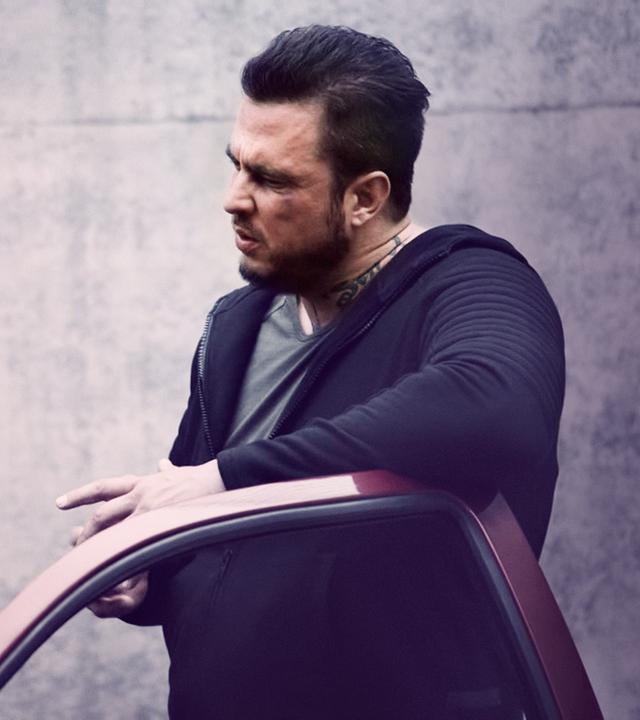 Peter (Dirk Heinrichs), ein Man mit einer schwarzen Lederjacke, blickt zu Dennis (Adrian Lynn) hoch, ein sehr großer und breiter Mann mit einem Tattoo am Hals. Die beiden stehen an einem Auto.