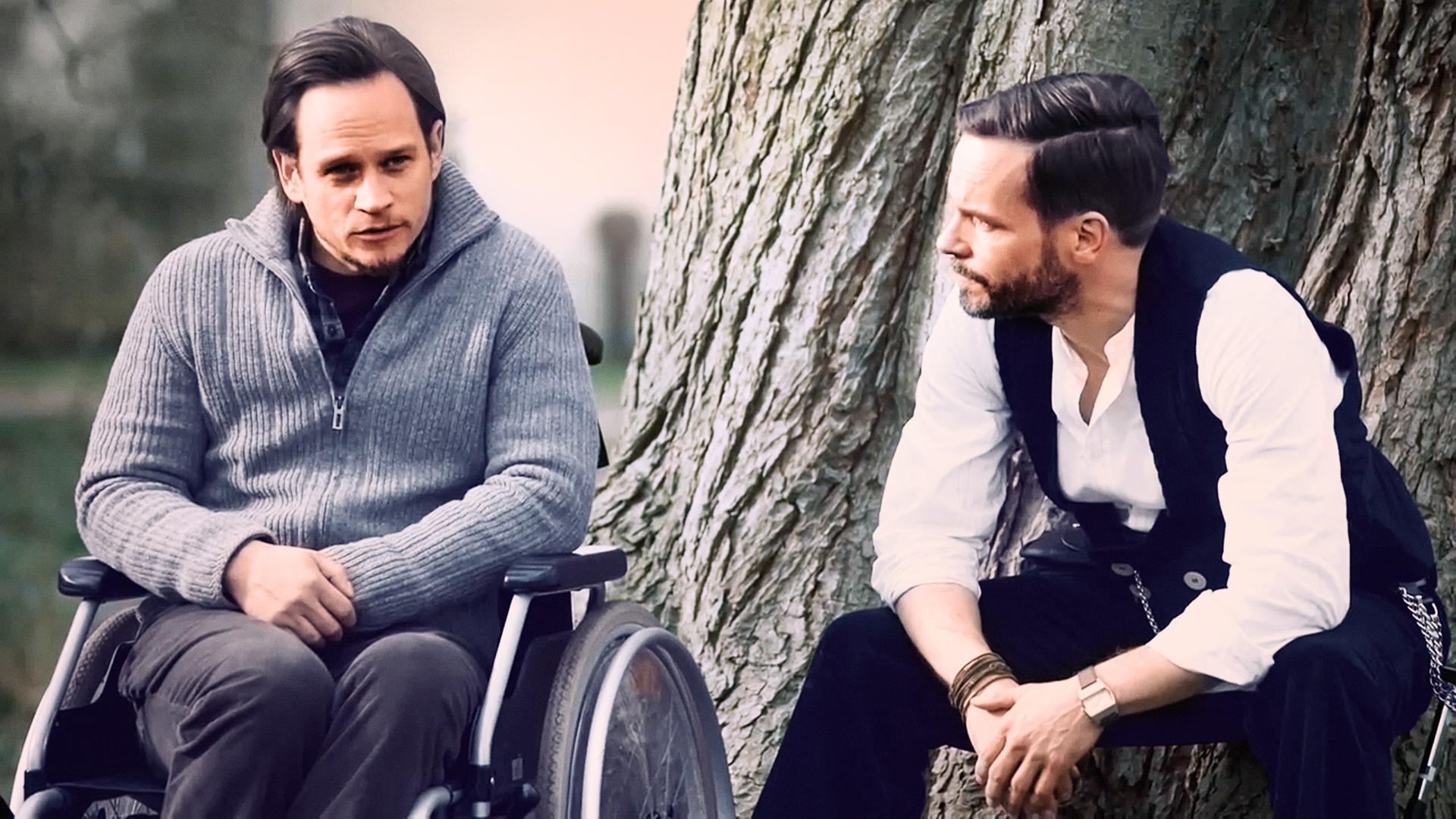 Leo Oswald (Wanja Mues) sitzt an einem Baum neben einen Mann (Vinzenz Kiefer), der in einem Rollstuhl sitzt.