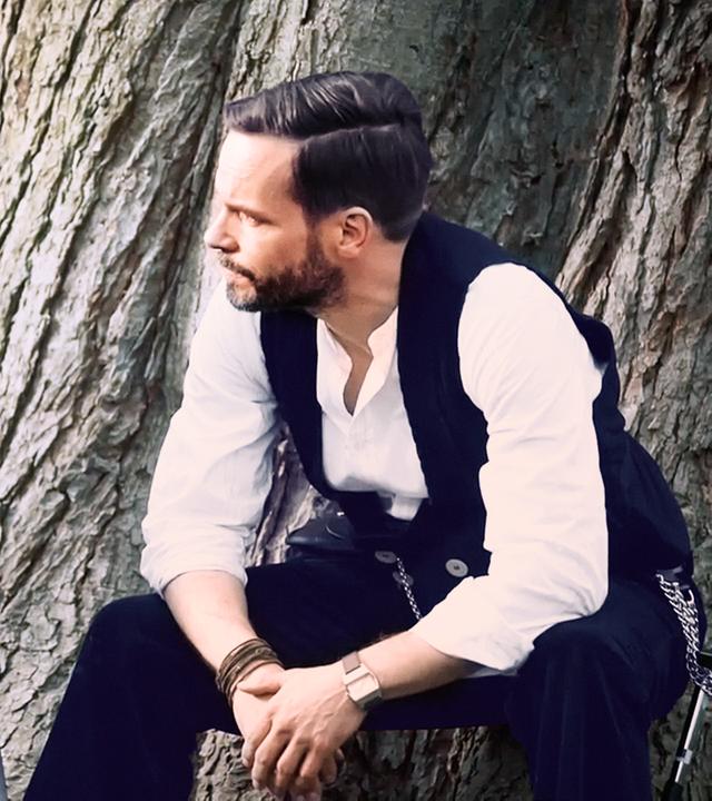 Leo Oswald (Wanja Mues) sitzt an einem Baum neben einen Mann (Vinzenz Kiefer), der in einem Rollstuhl sitzt.
